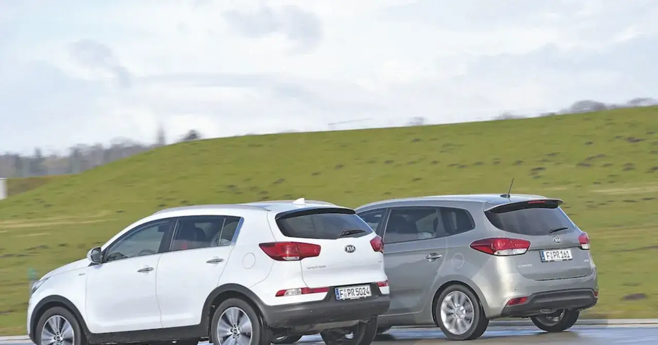 Biały Kia Sportage i srebrny Kia Carens 4 jadą obok siebie na tle zielonego wzgórza.