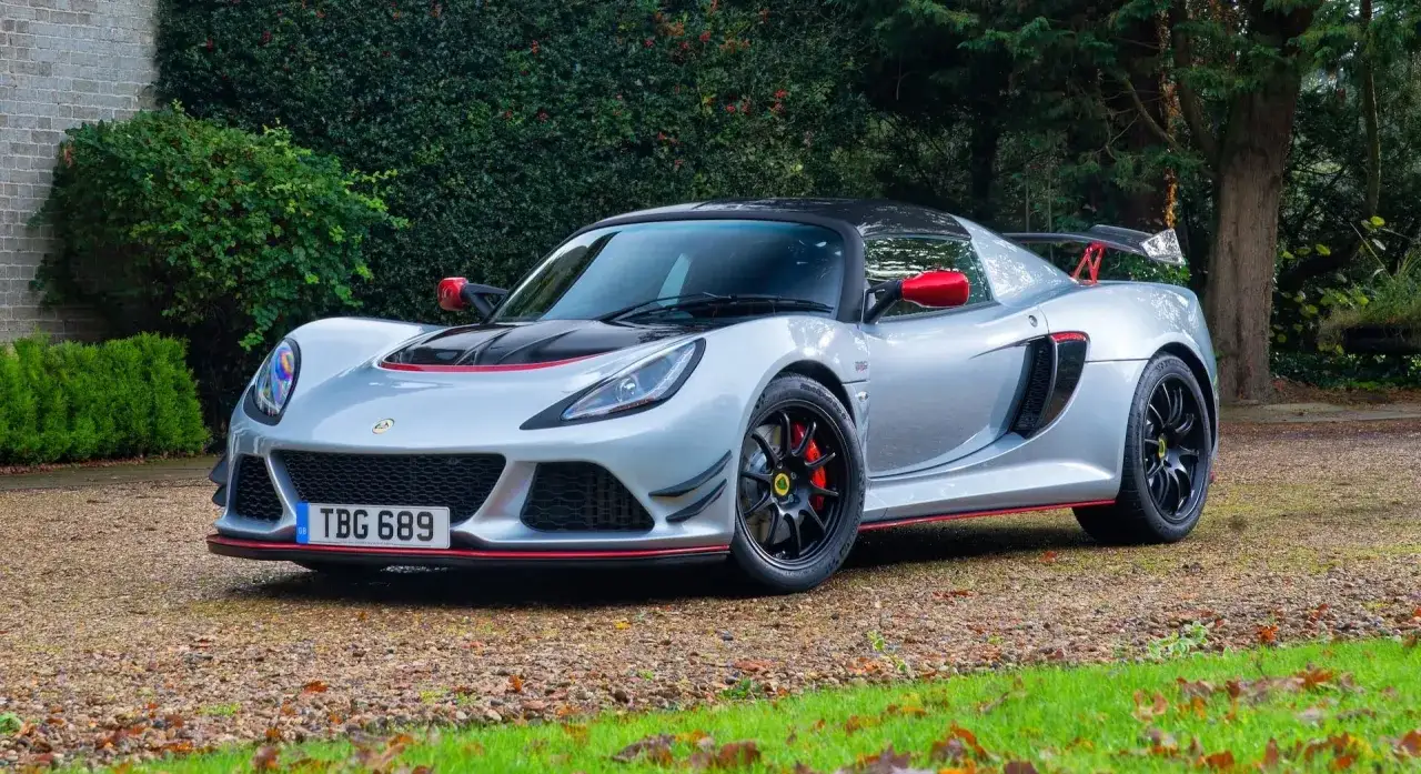 Srebrny Lotus Exige z czerwonymi akcentami i czarnym dachem, zaparkowany na żwirowej drodze, otoczony zielenią.