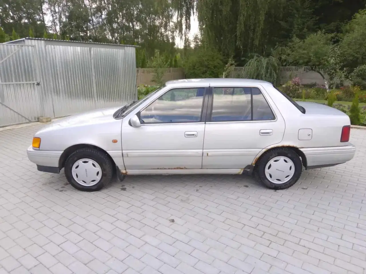 Srebrny Hyundai Pony z widocznymi śladami rdzy na progach, zaparkowany na kostce brukowej obok metalowego garażu.