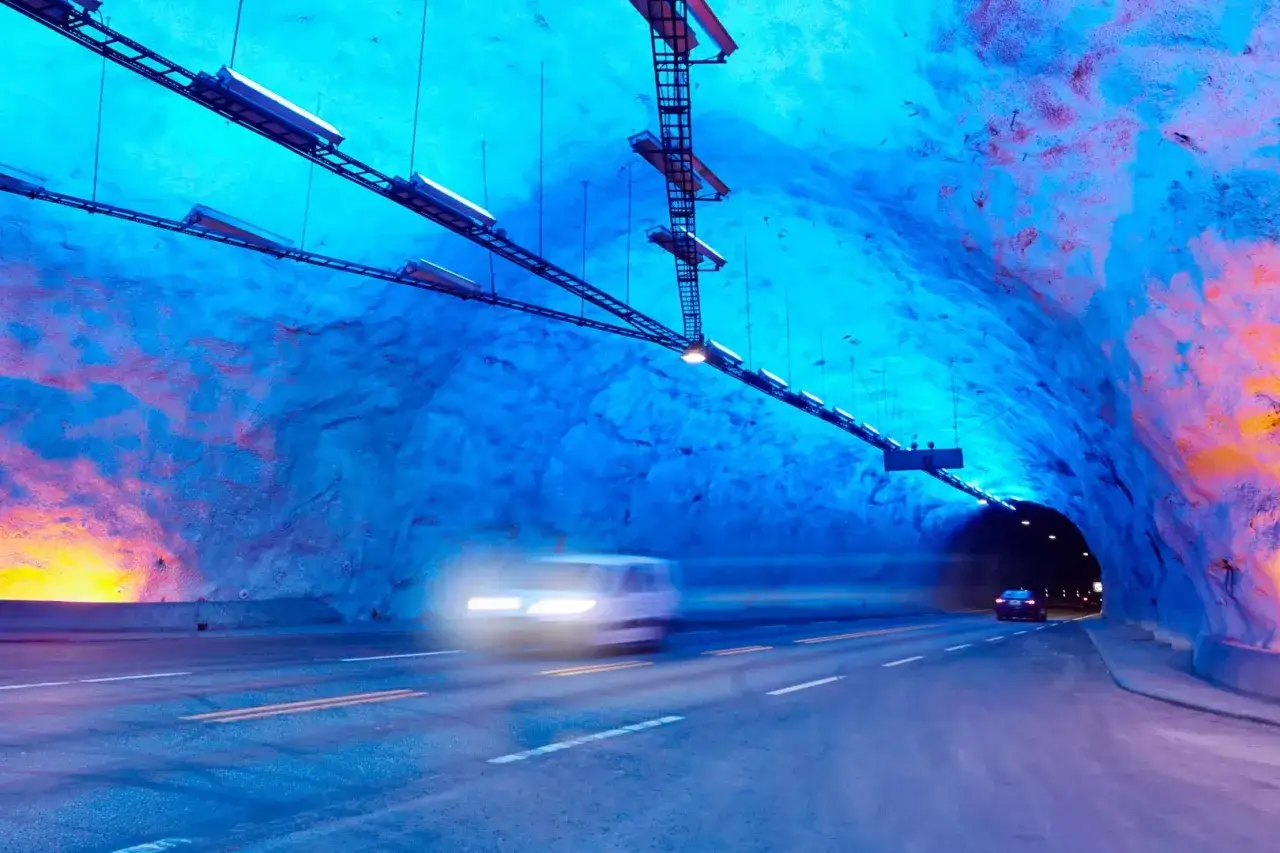 Wnętrze najdłuższego tunelu na świecie, oświetlone błękitnym i pomarańczowym światłem. Samochody pędzą w mrok.