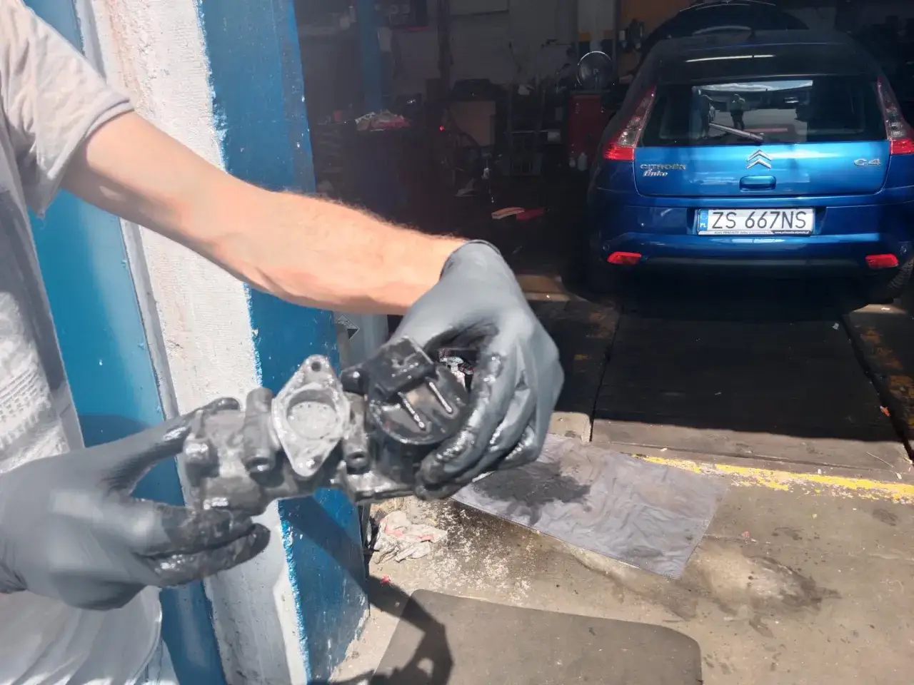 Mechanik w rękawiczkach trzyma brudny zawór EGR. W tle niebieski Citroen C4.