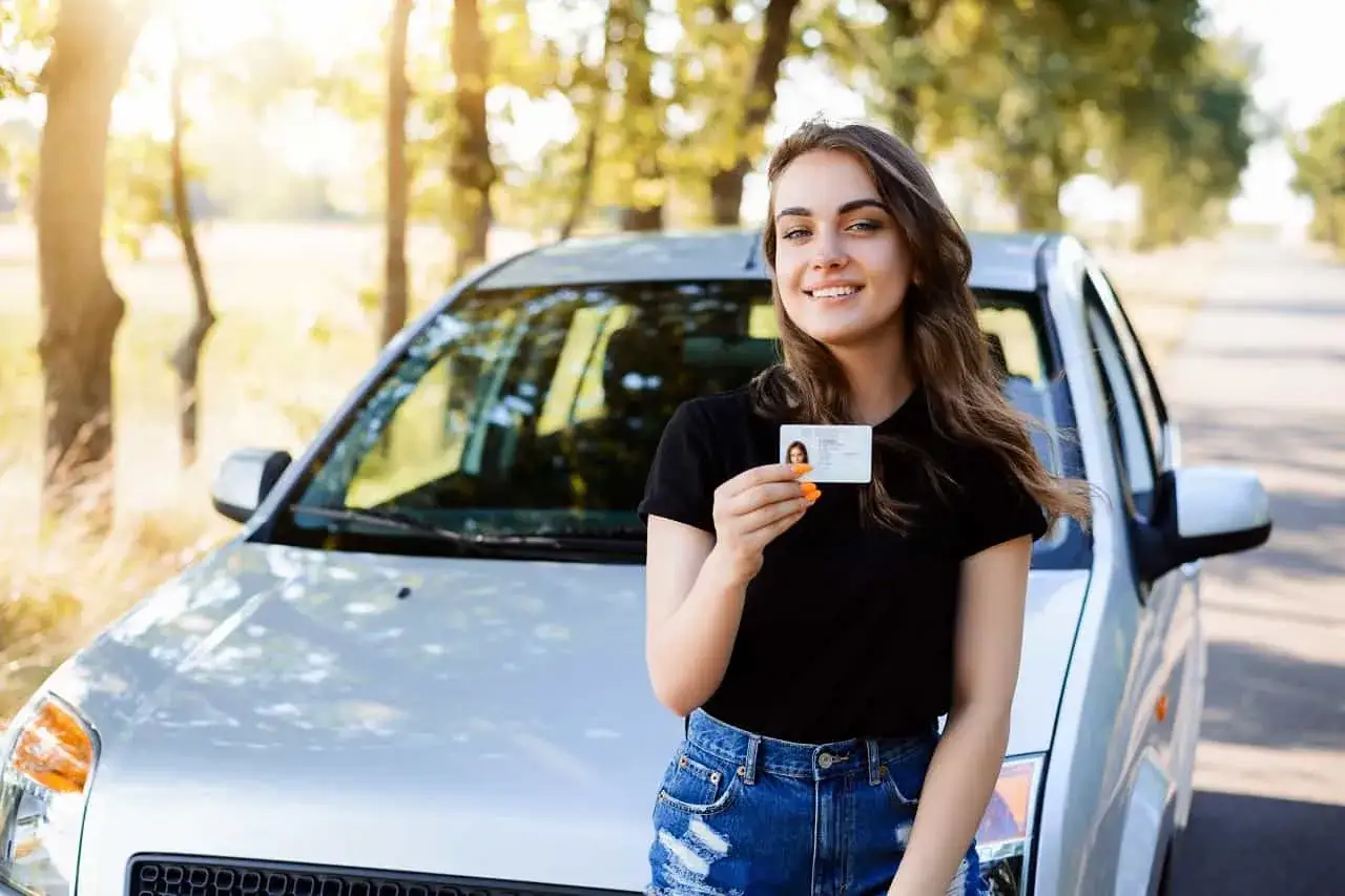Szczęśliwa młoda kobieta z prawem jazdy od 16 lat stoi obok samochodu na tle drzew.