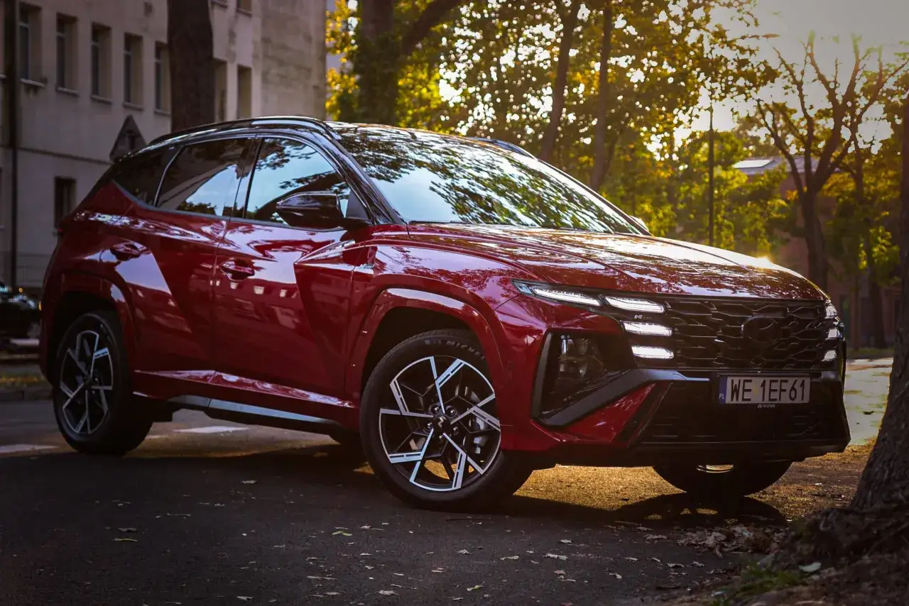 Czerwony Hyundai Tucson hybryda zaparkowany na tle jesiennych drzew i kamienicy.